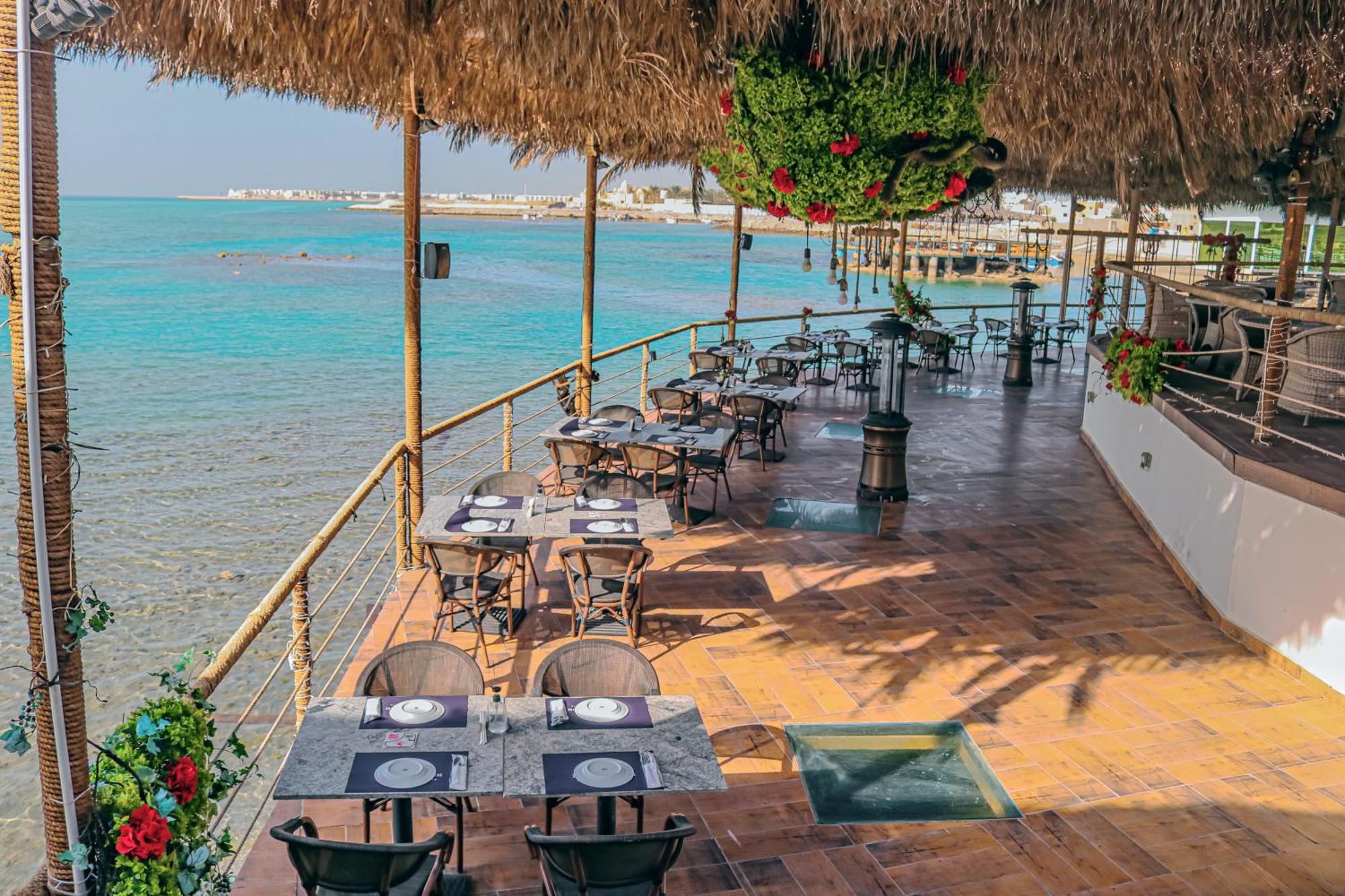 Lagoona Beach Luxury Resort And Spa Manama Exterior foto The photo shows a seaside restaurant with a spacious outdoor dining area. It features several tables set with plates and utensils, surrounded by lush greenery and decorative plants. The flooring is made of brown tiles, and there are thatched roofs pr