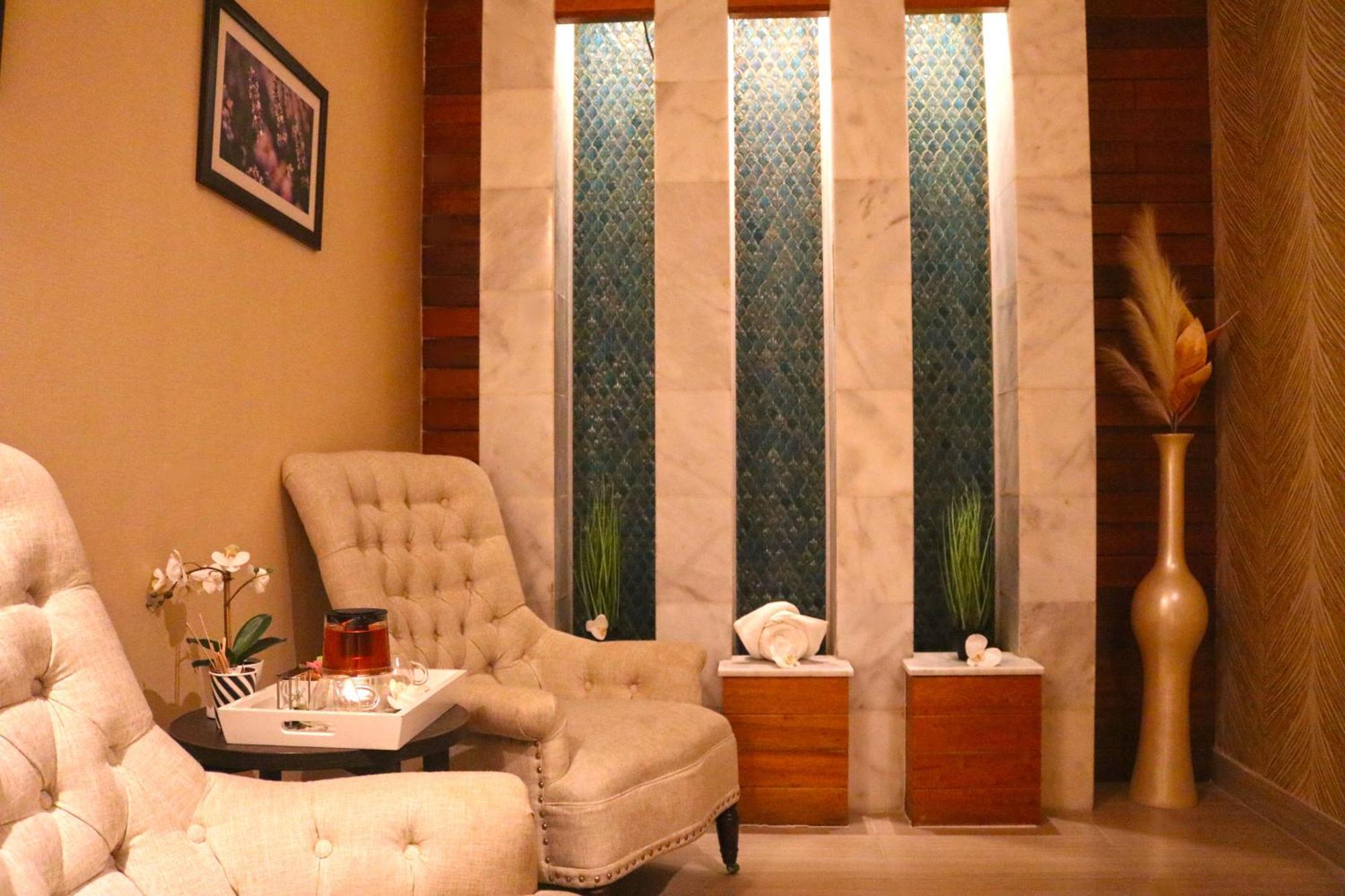 Lagoona Beach Luxury Resort And Spa Manama Exterior foto The image shows a relaxing interior space, likely a waiting area or lounge. There are two plush, beige armchairs positioned near a small table, which holds a glass pitcher and a cup. The background features tall, elegant windows with decorative blue 
