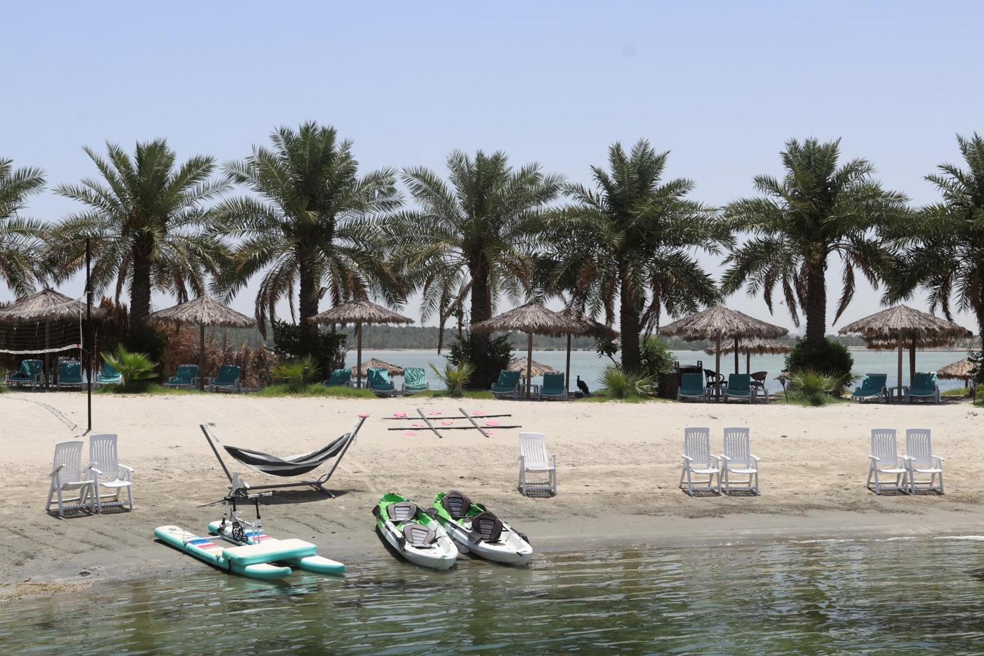Lagoona Beach Luxury Resort And Spa Manama Exterior foto The photo depicts a serene beach scene with several palm trees lining the shore. There are thatched umbrellas providing shade, and a few lounge chairs are positioned on the sand. A hammock is set up between two supports, inviting relaxation. In the f
