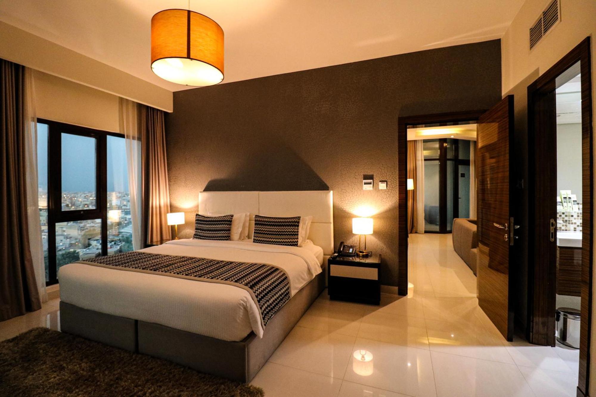 Lagoona Beach Luxury Resort And Spa Manama Exterior foto The photo depicts a modern hotel room or suite. It features a large bed with a white and gray color scheme, accented with decorative pillows. On either side of the bed, there are sleek bedside lamps providing warm lighting. The walls are a dark color