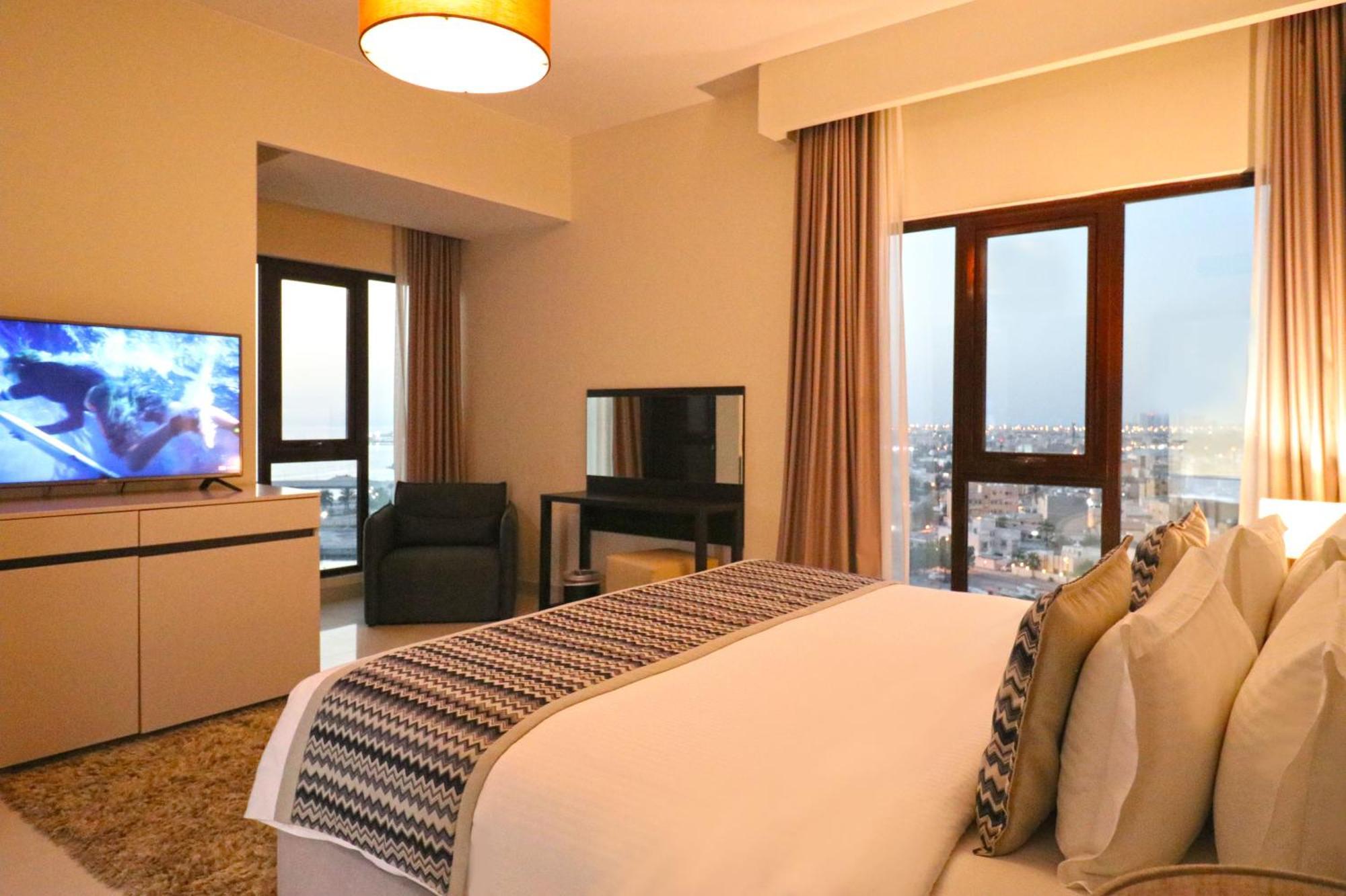 Lagoona Beach Luxury Resort And Spa Manama Exterior foto The photo shows a modern hotel room or suite. It features a neatly made bed with decorative pillows in the foreground. To the left, there is a TV on a cabinet, and further back, a comfortable chair is situated in front of a large window that likely o