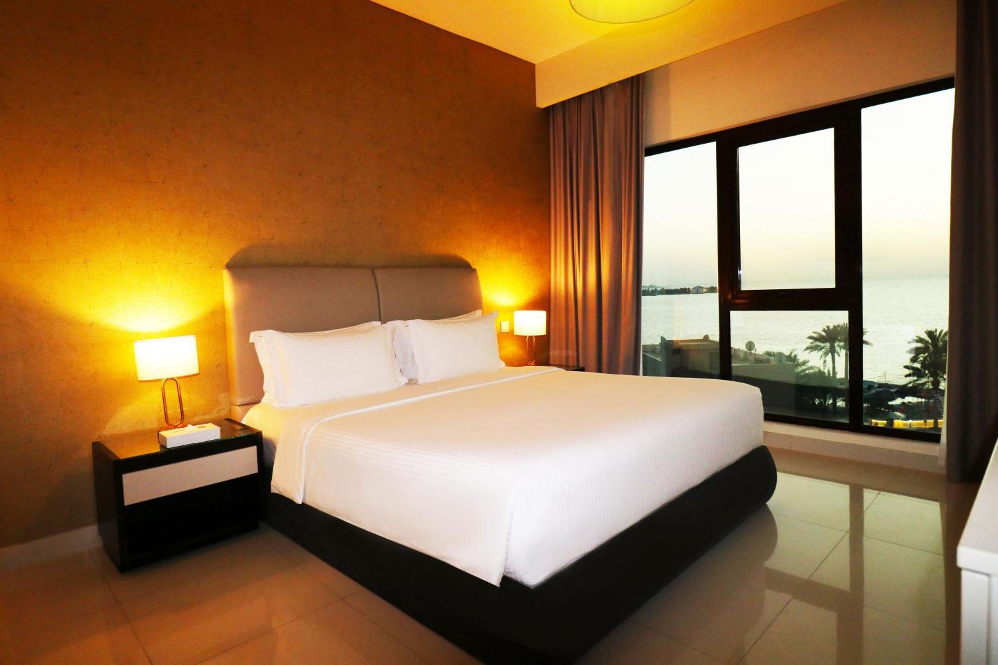 Lagoona Beach Luxury Resort And Spa Manama Exterior foto The photo shows a modern hotel bedroom with a large, comfortable bed dressed in white linens. The room features warm lighting, including bedside lamps, creating a cozy atmosphere. There are large windows with a view of the outdoors, possibly overlook